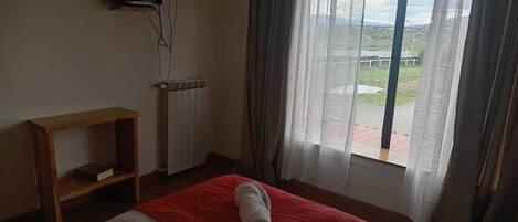 Habitación de hotel con cama, televisión montada en la pared, radiador y ventana con cortinas.