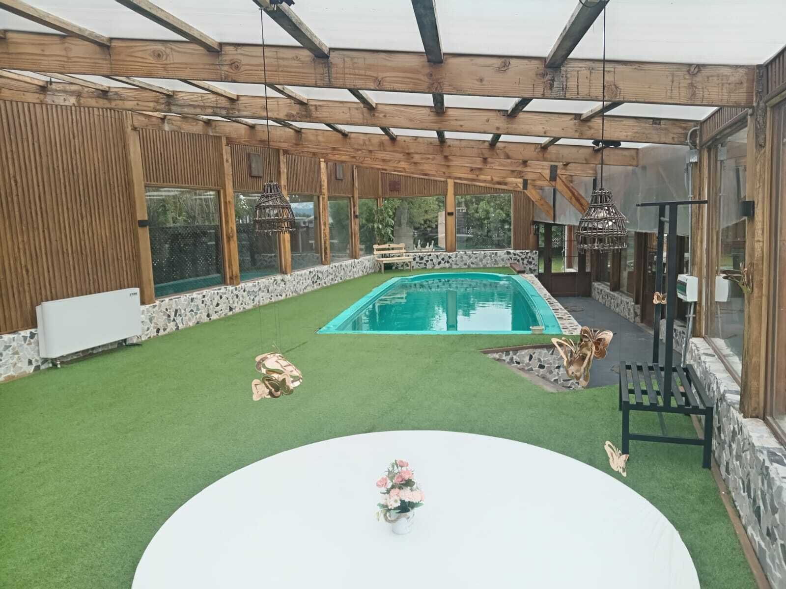 A spacious indoor pool area with a wooden ceiling and stone walls, surrounded by artificial grass and a central white table with a floral arrangement.