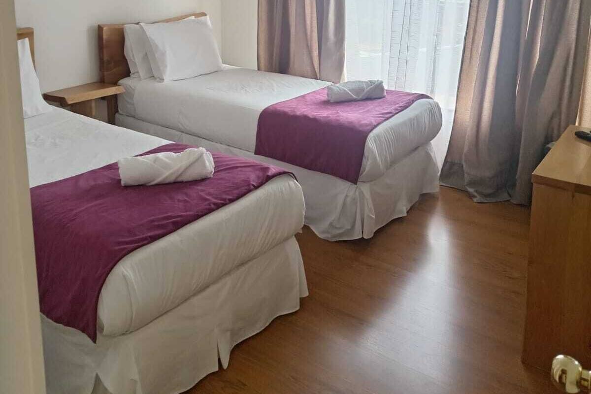 Habitación de hotel con dos camas, una mesita de noche de madera, un escritorio y una ventana con cortinas.