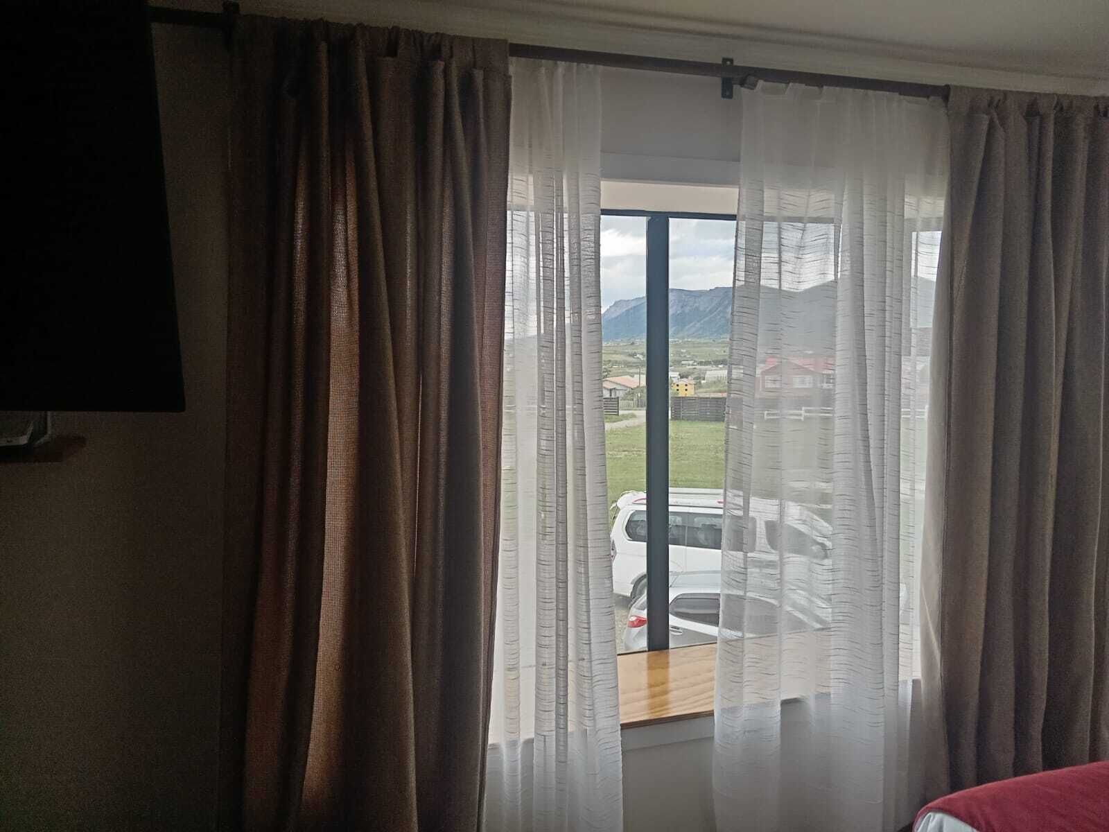 A room with a window featuring sheer curtains, a view of a white car outside, and a red bed in the foreground.