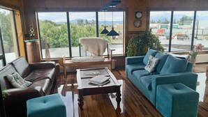 Een gezellige woonkamer met houten vloer, een bruine leren bank, een blauwgroene bank en een houten salontafel. Er zijn grote ramen met uitzicht op groen en bergen.