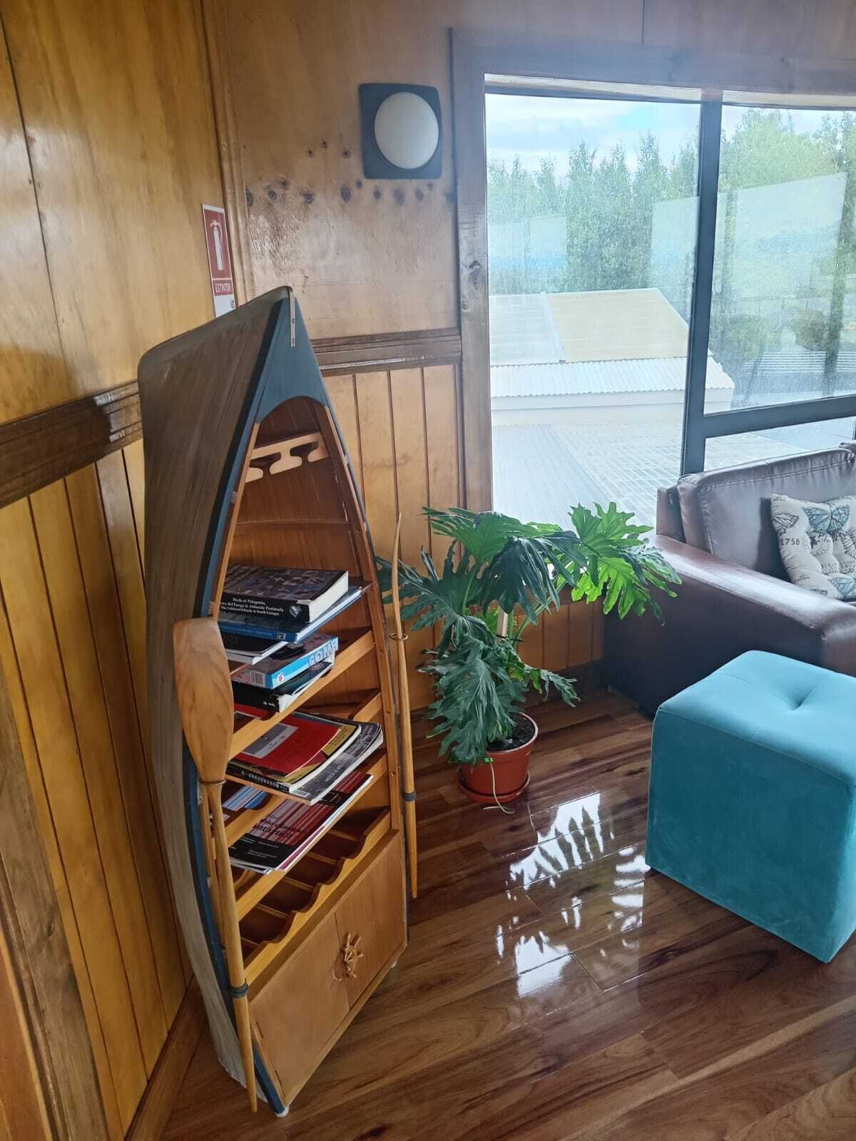 Una estantería con forma de barco de madera llena de libros, una planta en maceta, un reposapiés azul y una ventana con vistas a un edificio y árboles.