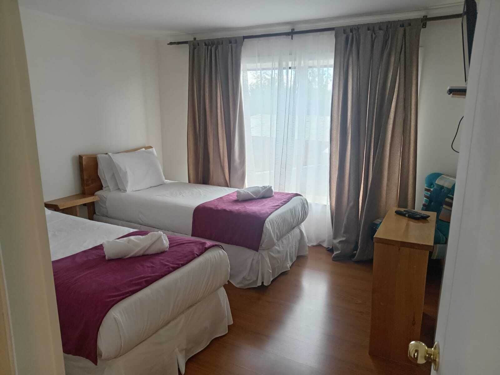 A hotel room with two beds, a wooden desk, and a view through sheer curtains.