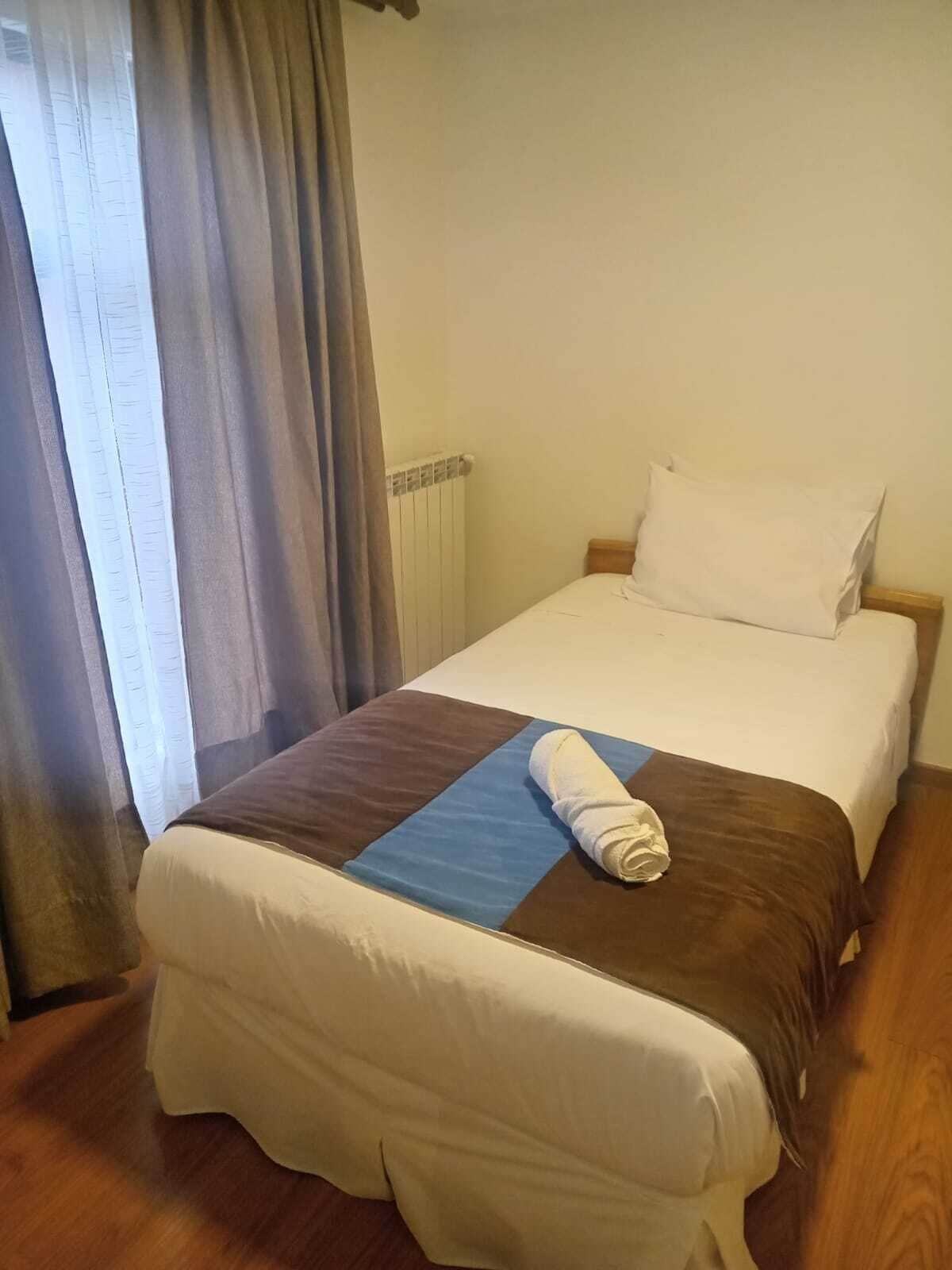 A single bed with a brown and blue bedspread, white pillows, and a rolled towel on the bed.