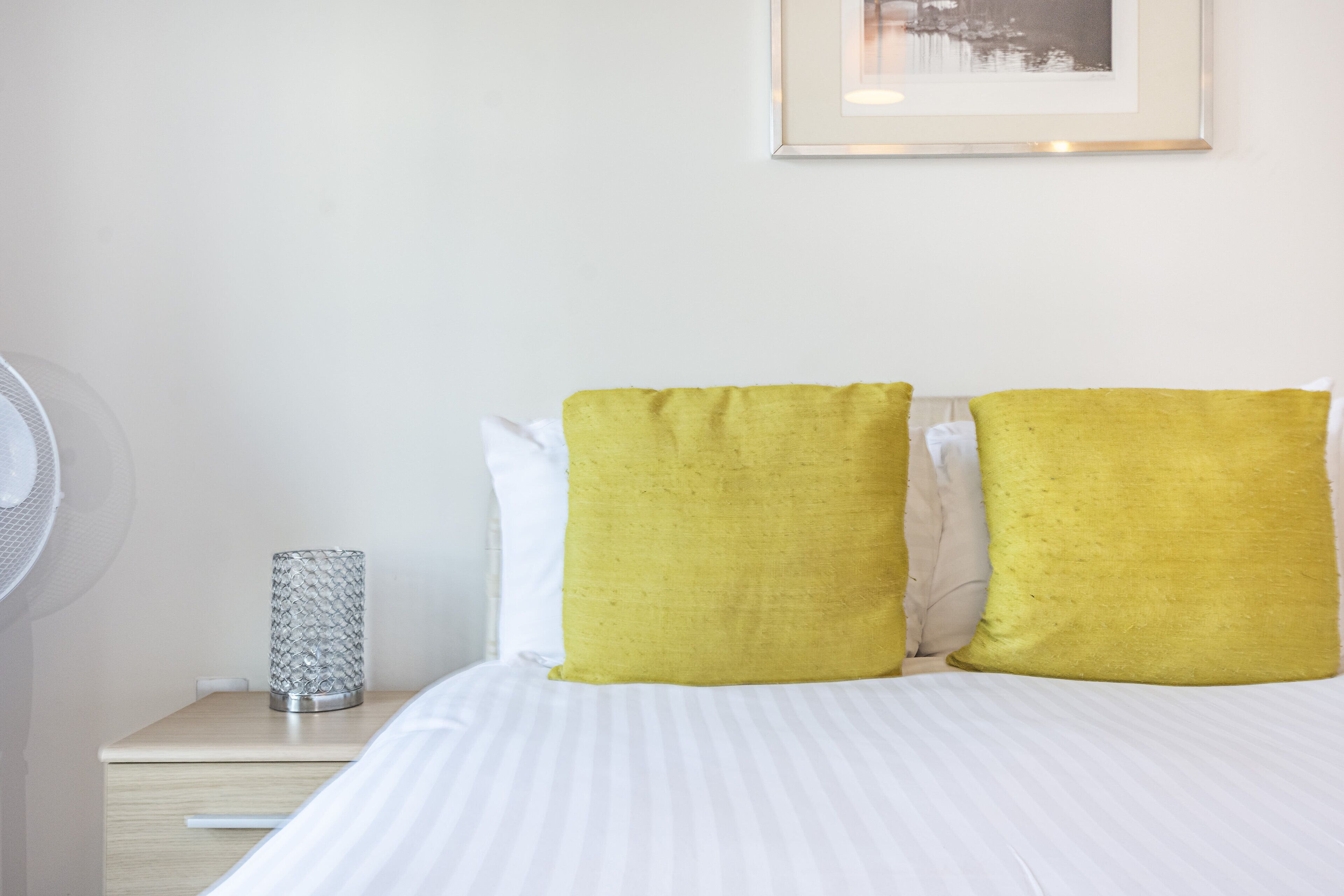 Un letto rifatto con biancheria bianca e due cuscini gialli, un comodino con una lampada e un quadro appeso al muro.