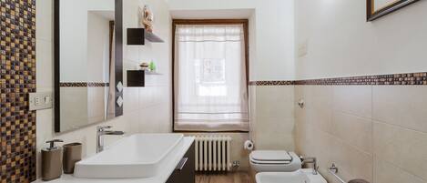 Casa de banho com uma banheira grande, sanita, lavatório e janela com cortinas.