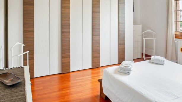 A hotel room with a wooden floor, a bed with white linens, a ceiling fan, and a window with curtains.
