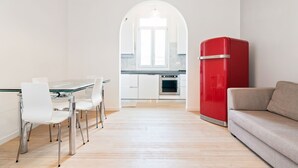 Une cuisine moderne avec des meubles blancs, un réfrigérateur rouge et un coin repas comprenant une table en verre et des chaises blanches.