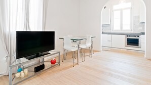 Un salon moderne avec un téléviseur à écran plat posé sur une table en verre, des chaises blanches et un coin cuisine doté de meubles blancs et d’un four.