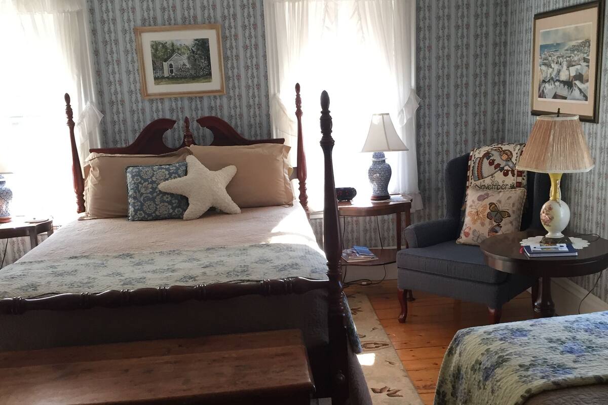 A bedroom with a wooden four-poster bed, a blue armchair, a small round table with a lamp, and a nightstand with a lamp.