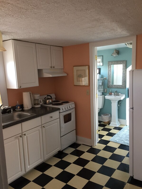 A kitchen with white cabinets, a stove, and a sink. There is a checkered floor and a doorway leading to a bathroom with a sink and mirror. - Lantana House (Rockport)