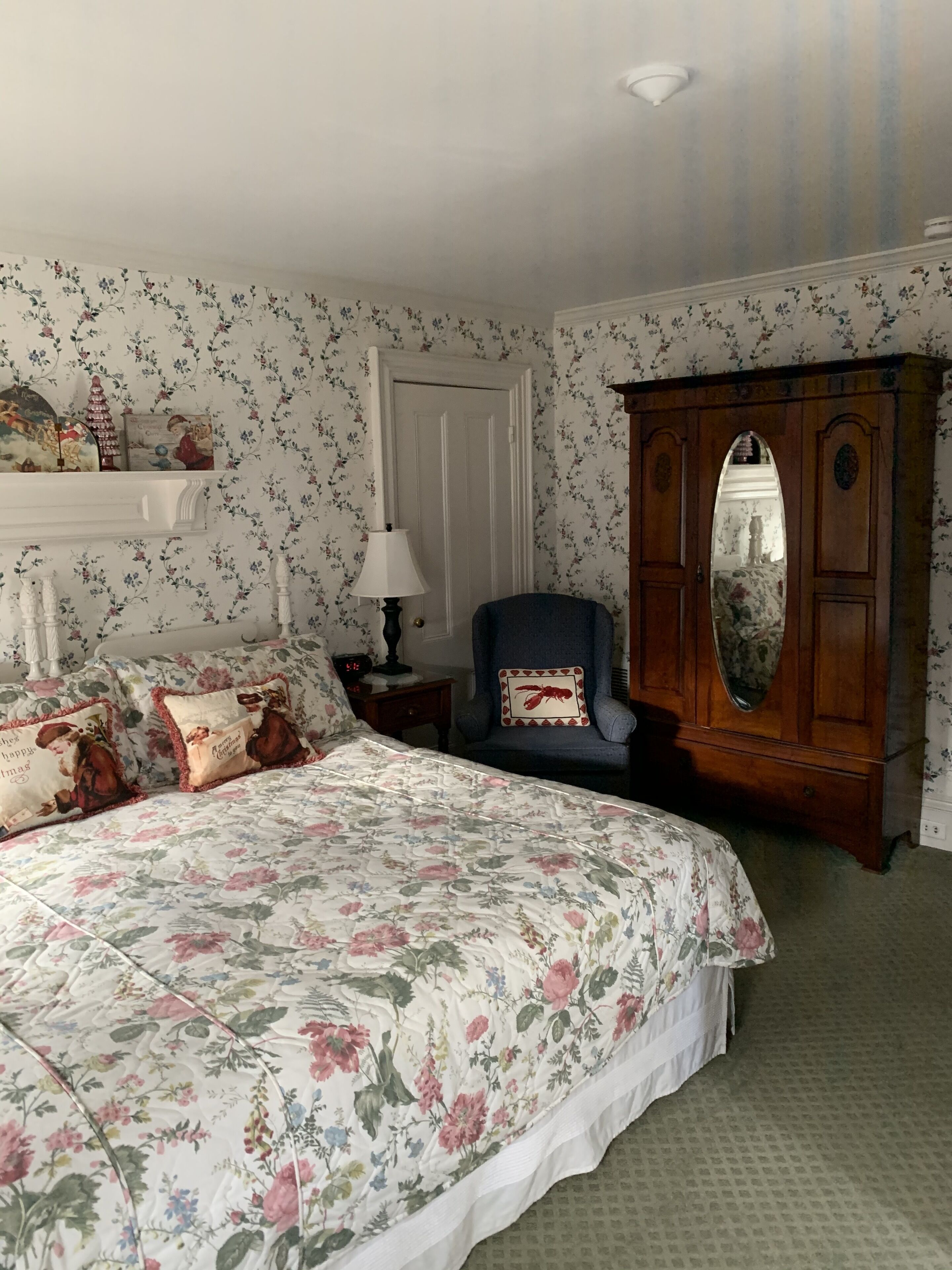 A bedroom with floral wallpaper, a bed with a floral bedspread, a wooden wardrobe, and a nightstand with a lamp.