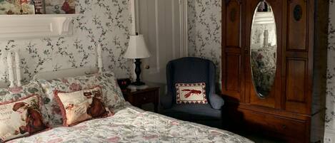 A bedroom with floral wallpaper, a bed with a floral bedspread, a wooden wardrobe, and a nightstand with a lamp.