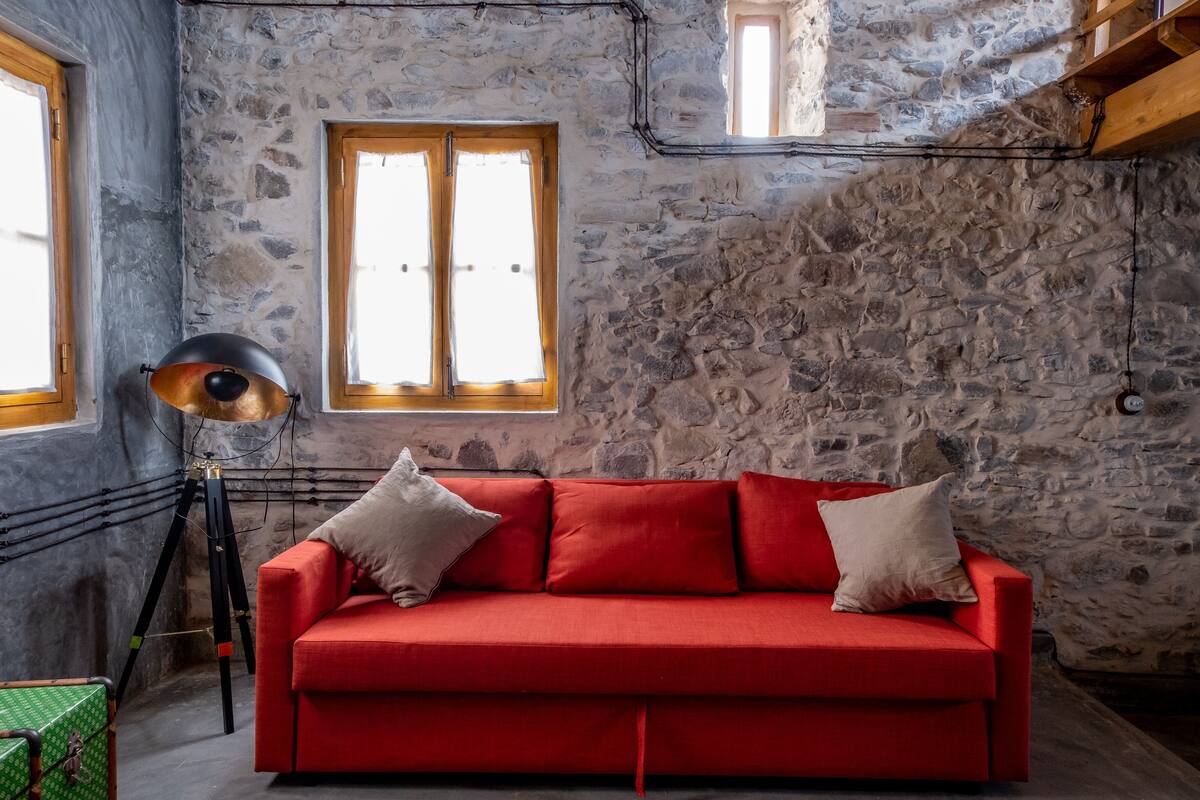 A red sofa with two pillows, a tripod lamp, and a stone wall with a window.