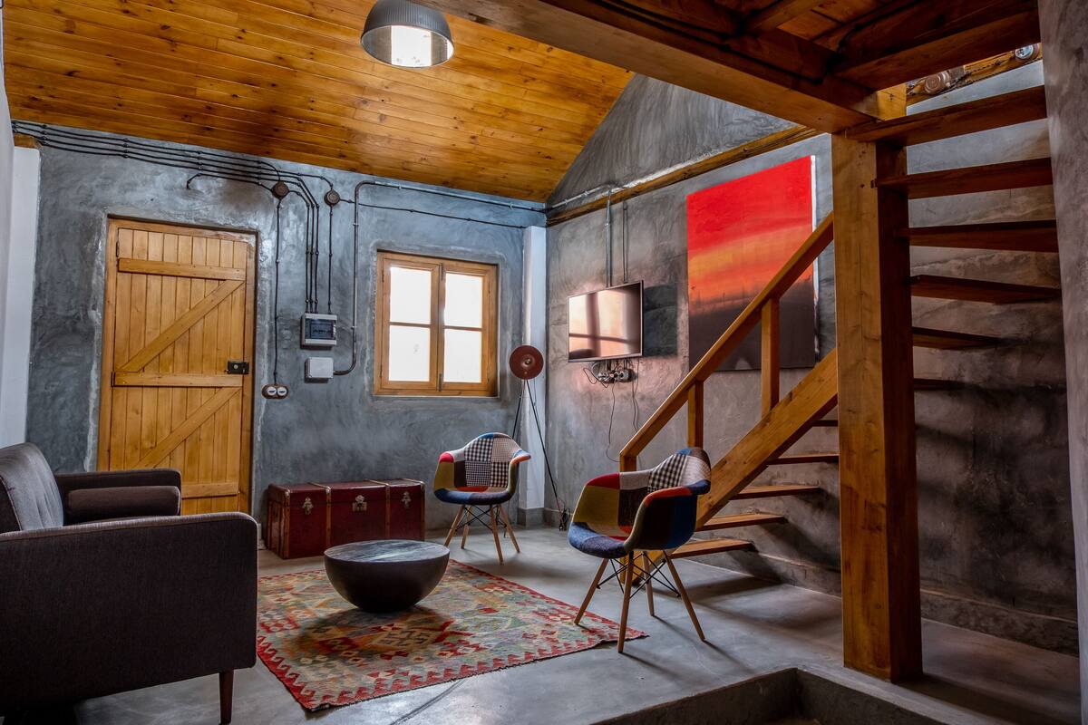 A rustic room with a wooden ceiling, a large window, a wooden door, a TV, a fireplace, and a staircase.
