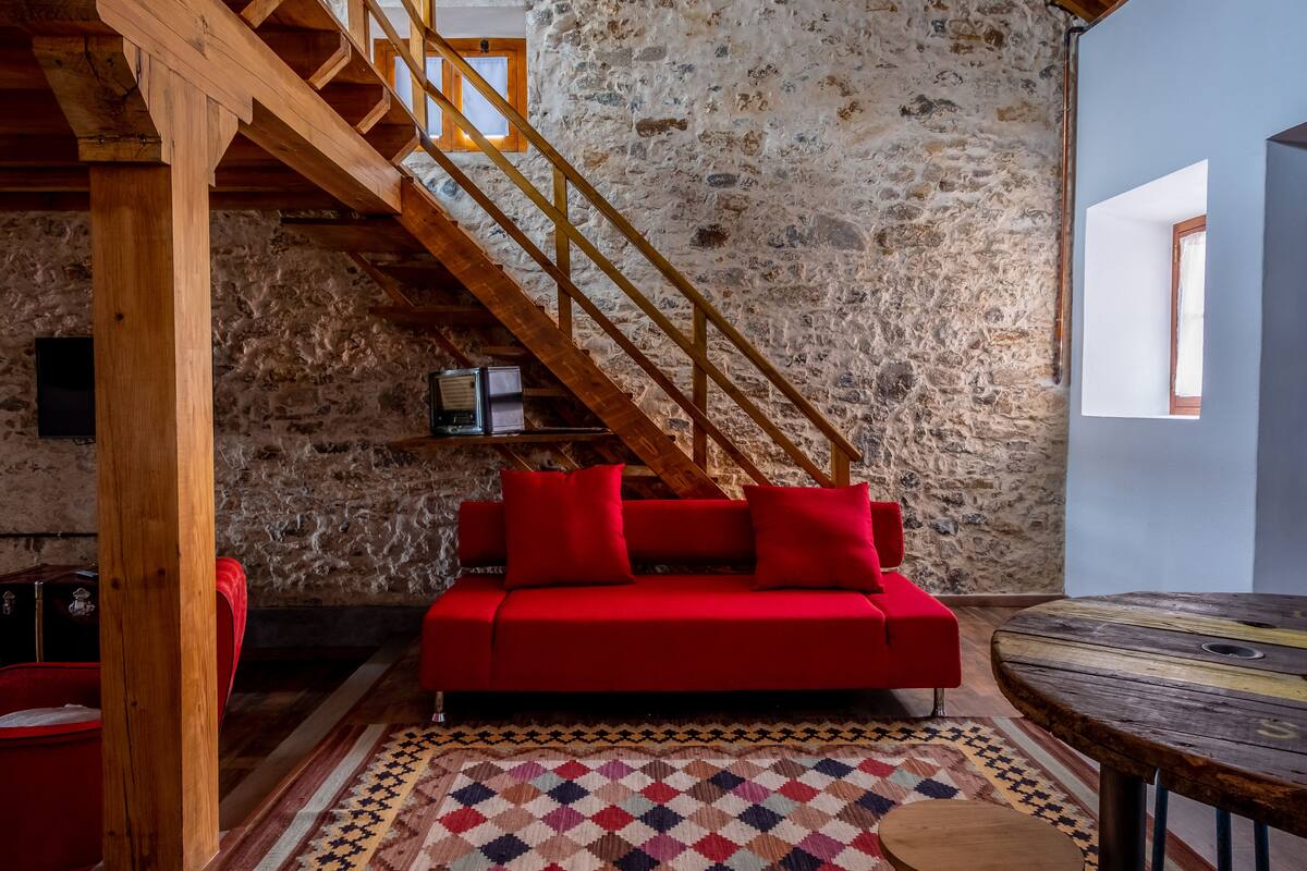 A cozy room with a red sofa, a wooden staircase, and a stone wall.