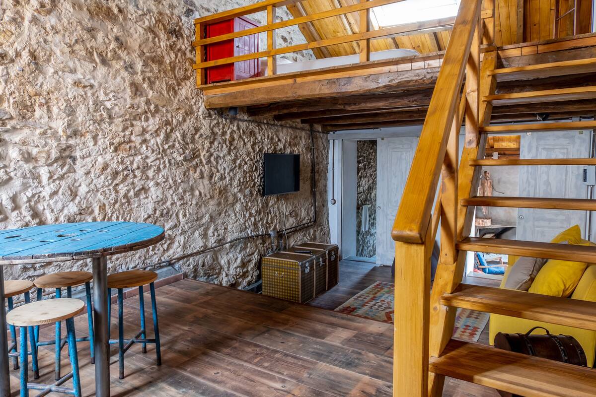 A rustic interior with a wooden staircase, a stone wall, a round table with stools, and a door leading to another room.