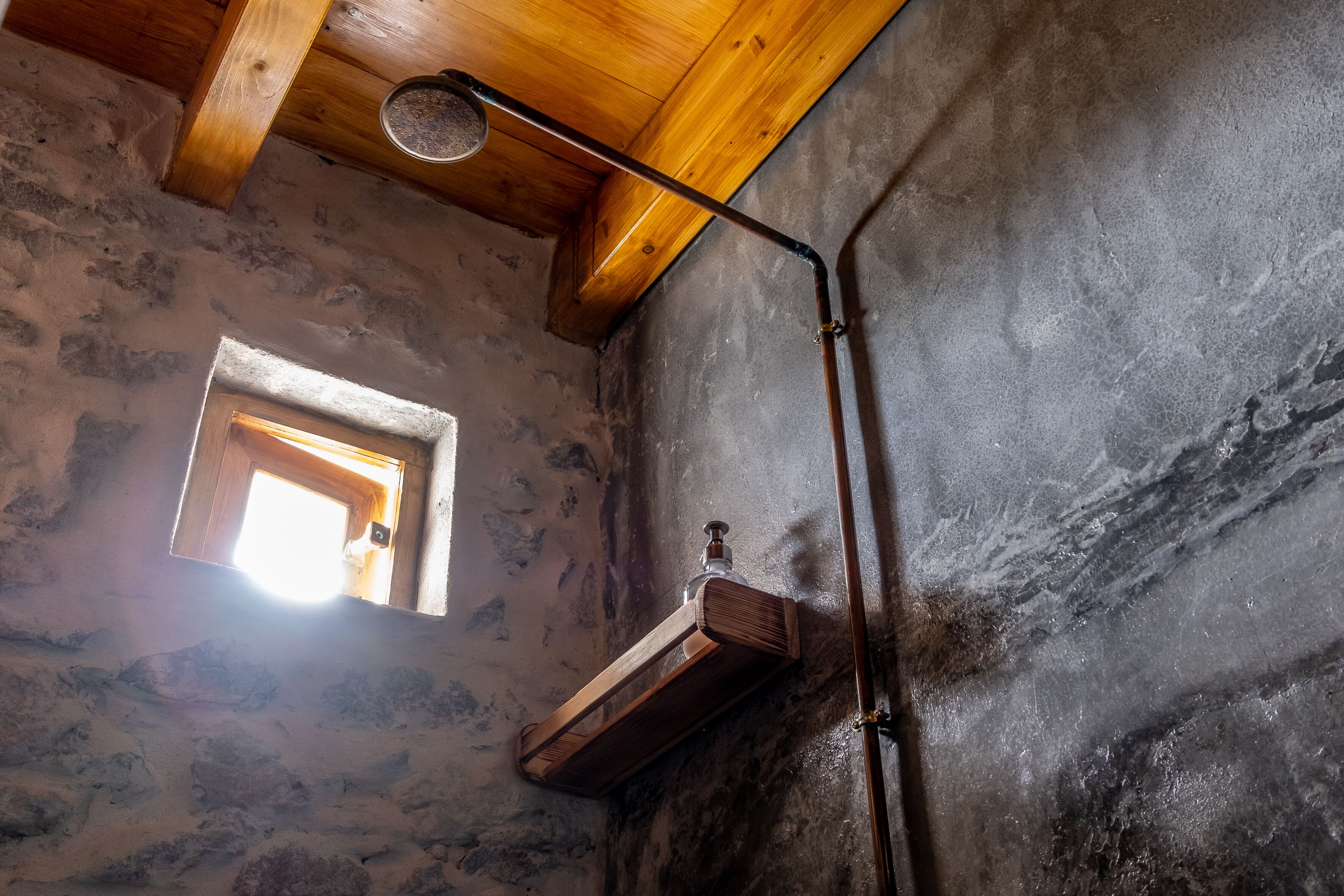 A rustic room with a wooden beam ceiling, a small window, and a metal pipe fixture.