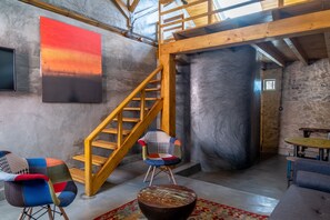 A room with a wooden staircase, a large abstract painting, a colorful patchwork chair, and a rustic wooden table. - Terra Lodge (Mafra)