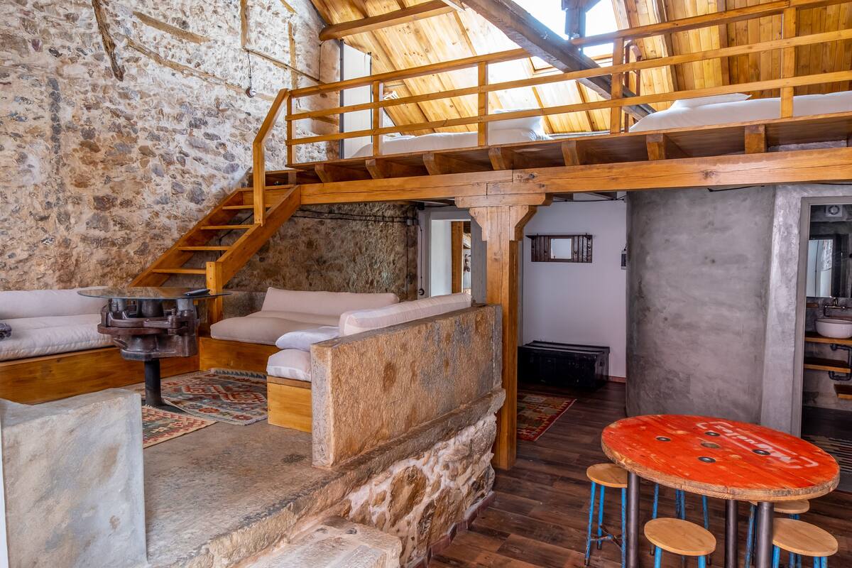 A rustic interior with a stone wall, wooden staircase, and a loft bed.