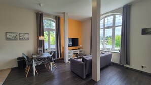 A modern living room with a dining area, large windows, and a flat-screen TV. - Apartments Villa Ratskopf (Wernigerode)