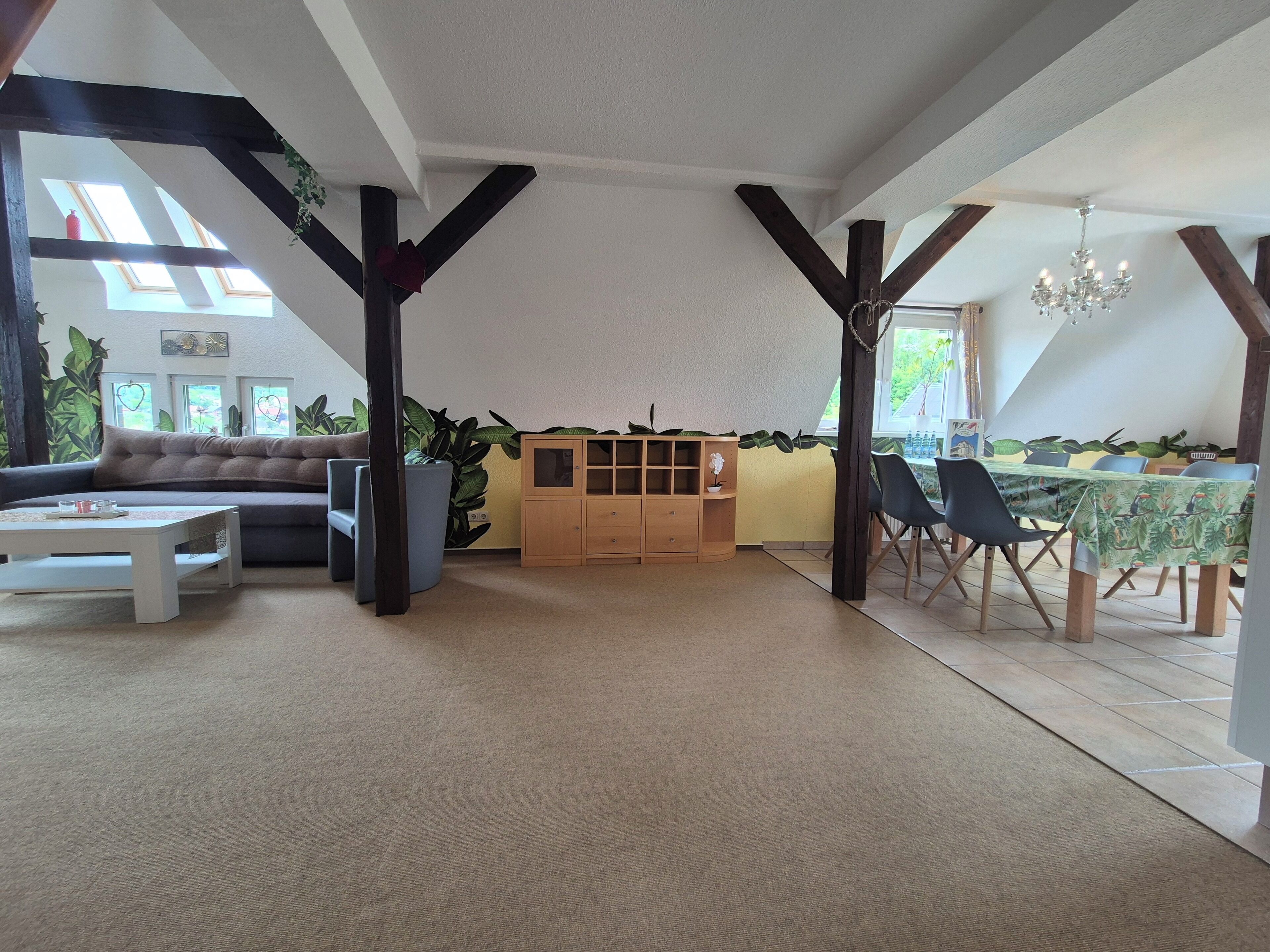 A spacious living area with a sofa, coffee table, and a wooden cabinet. There is a dining area with a table and chairs. The room has a sloped ceiling with exposed beams and large windows allowing natural light.