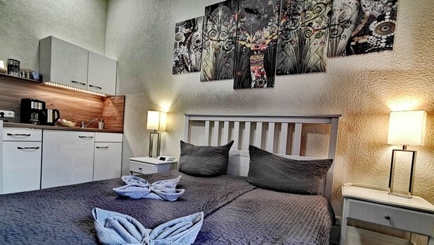 A hotel room with a bed, two bedside lamps, a nightstand, a small table, a coffee maker, and a kitchen area with cabinets and a sink. - Apartments Villa Ratskopf (Wernigerode)