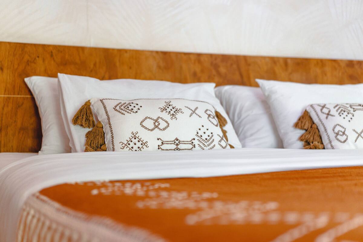 A bed with white pillows featuring embroidered designs and tassels, a wooden headboard, and an orange blanket.