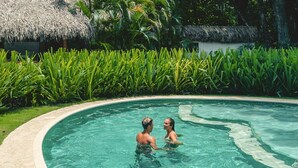 Una piscina con borde curvo, rodeada de exuberante follaje verde y una estructura con techo de paja al fondo.