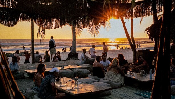 Zona de estar frente a la playa, con mesas de piedra y cojines, con vista al océano al atardecer.