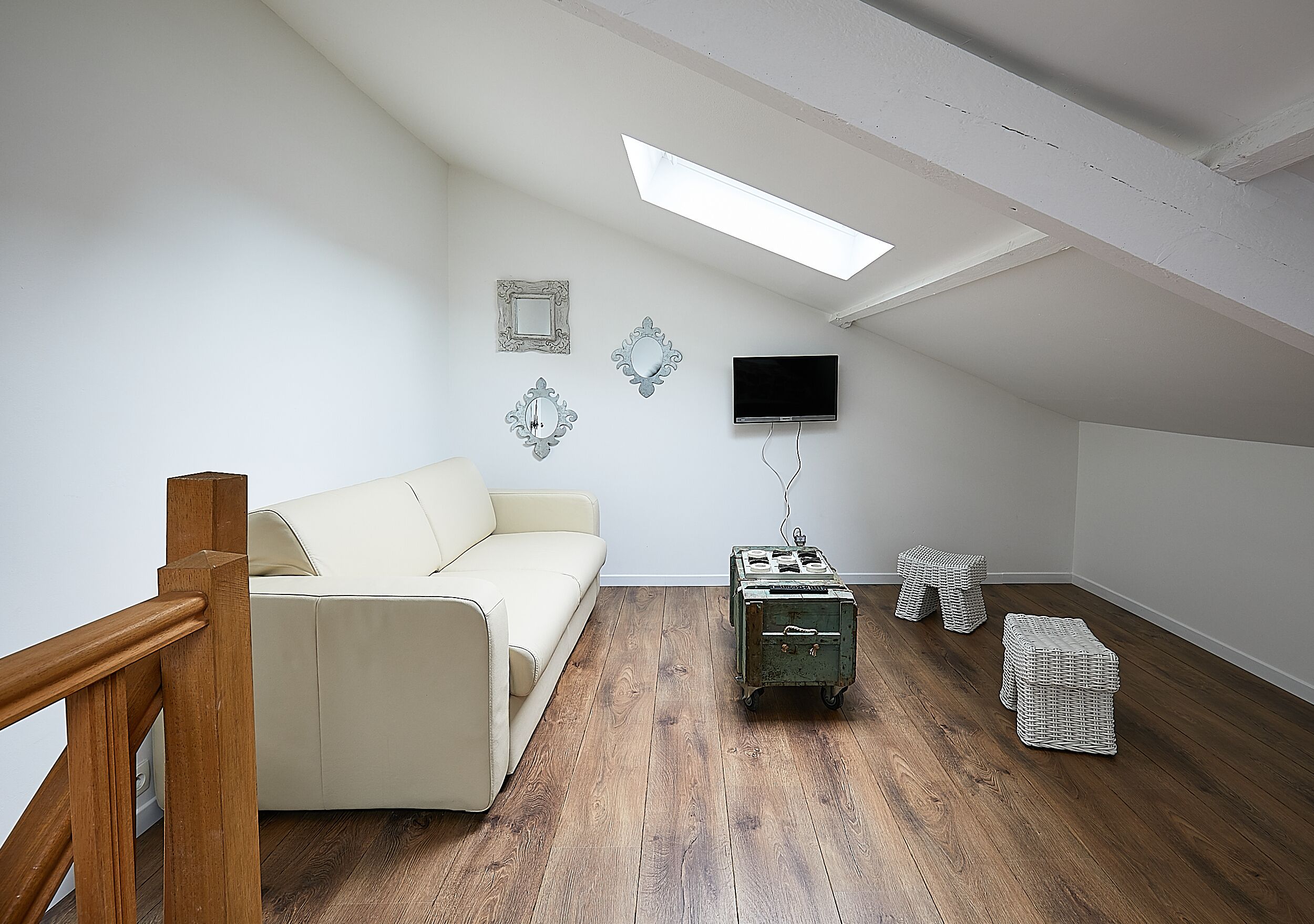 A room with a wooden floor, a white sofa, a television mounted on the wall, and a skylight.