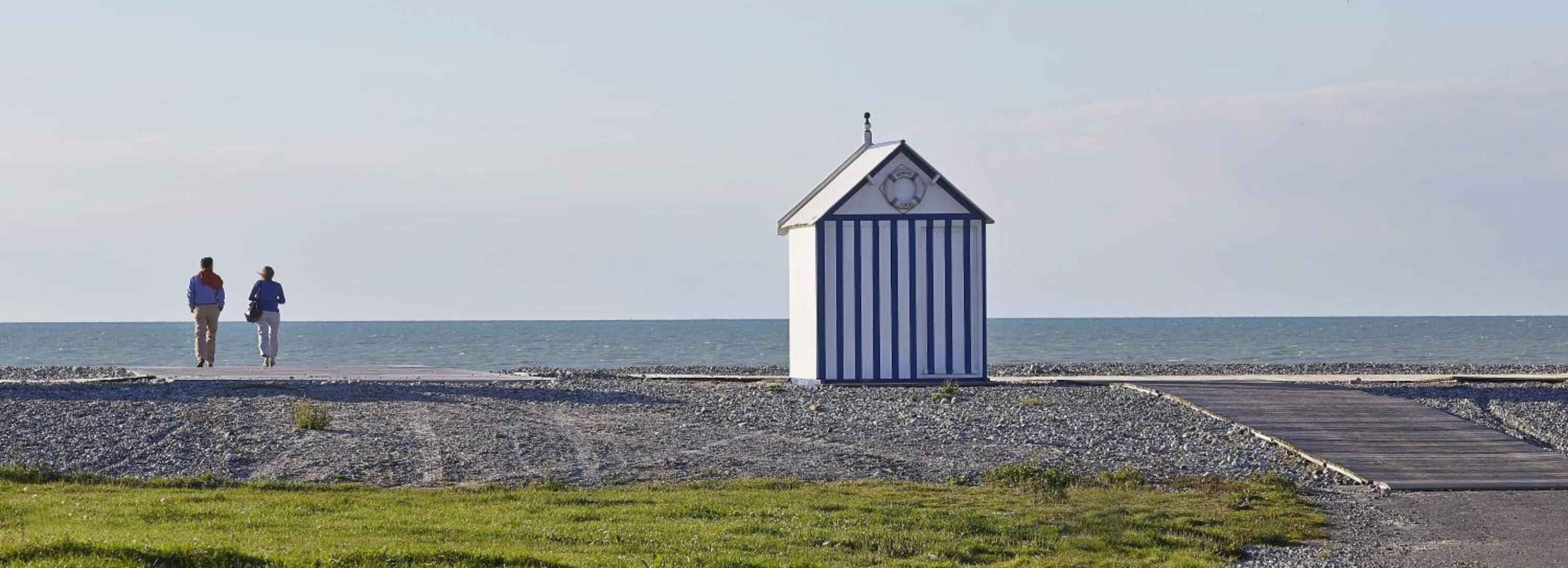 A beach with a small blue and white striped building, a wooden boardwalk, and two people walking.