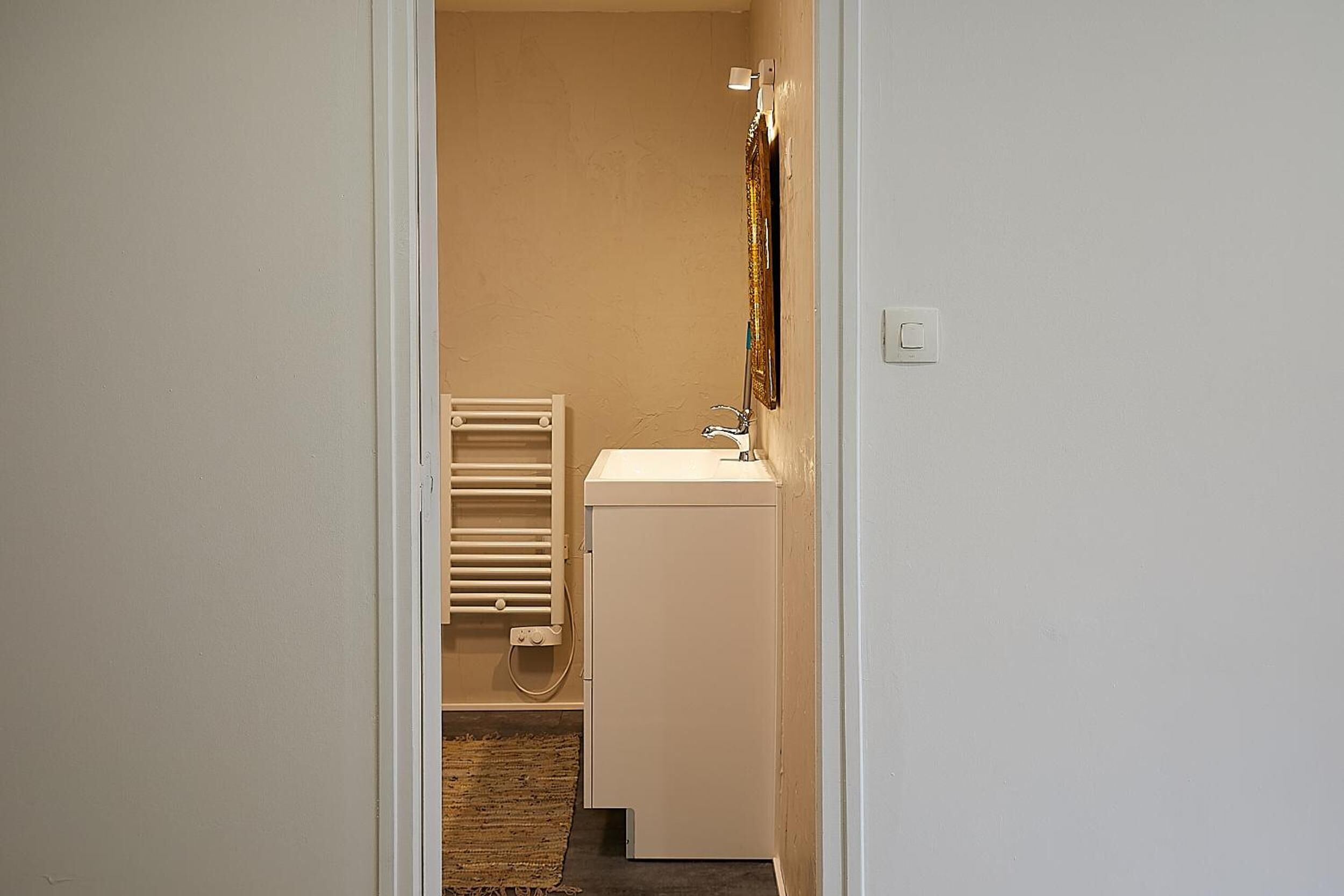 A narrow hallway leading to a bathroom with a white sink, a towel rack, and a radiator.