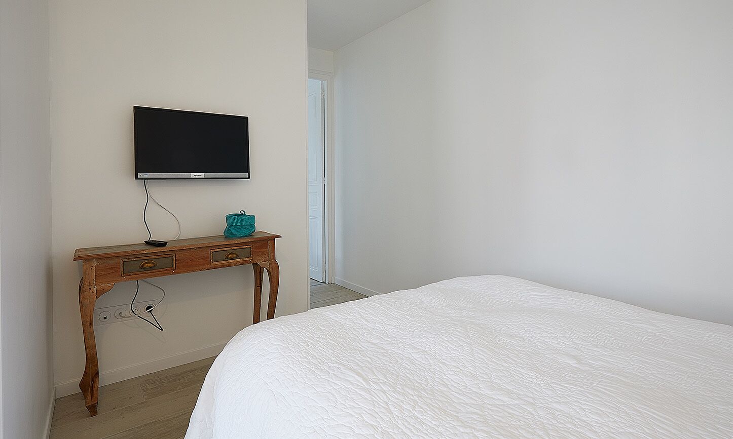 Une chambre à coucher avec un lit, un bureau en bois avec une télévision, et une porte donnant sur une autre pièce.