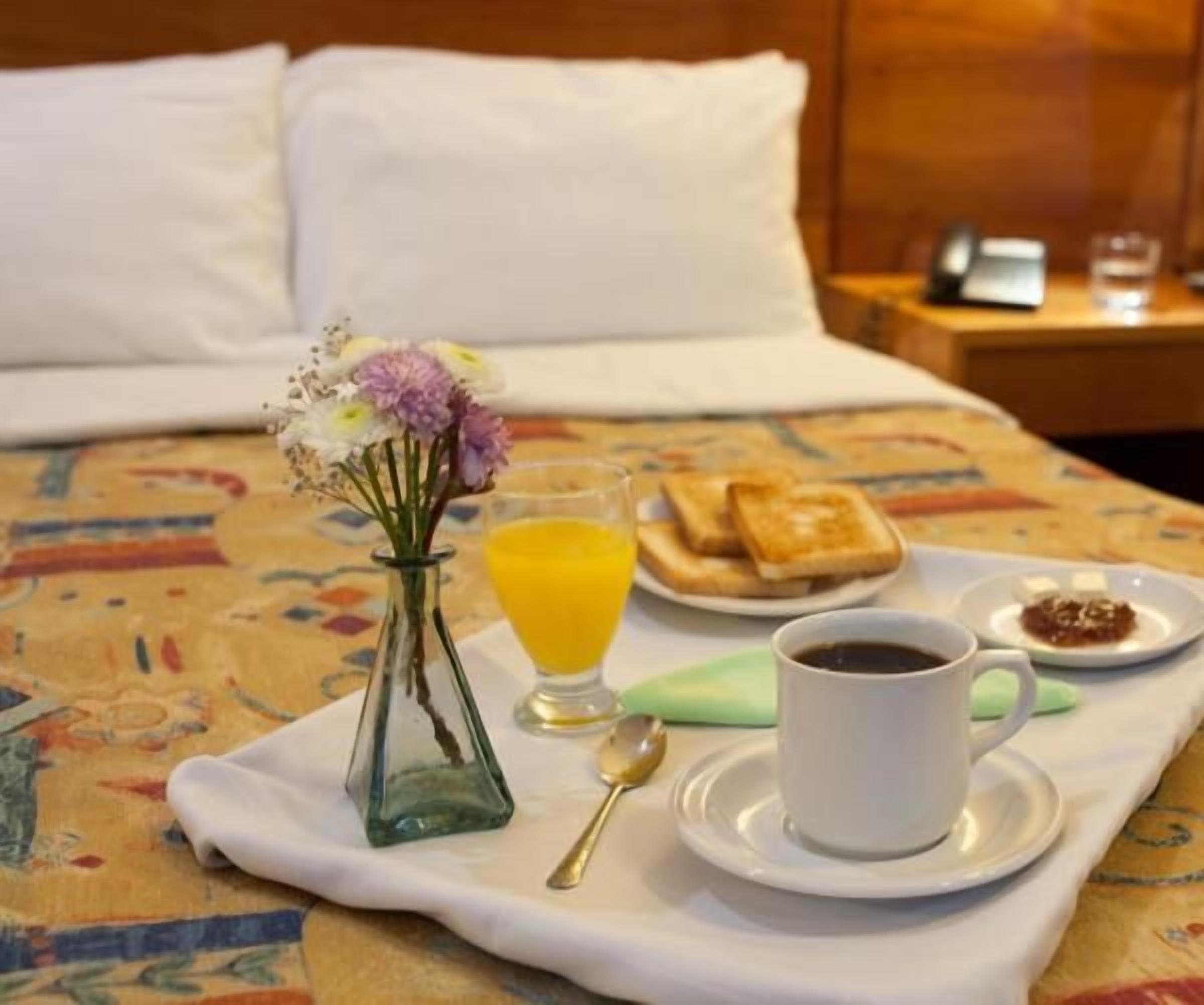 A neatly made breakfast tray on a bed with a floral vase, orange juice, toast, jam, and a cup of coffee.