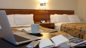 Una habitación de hotel con cama, una mesita de noche con lámpara, una computadora portátil, una taza, un control remoto y un libro.
