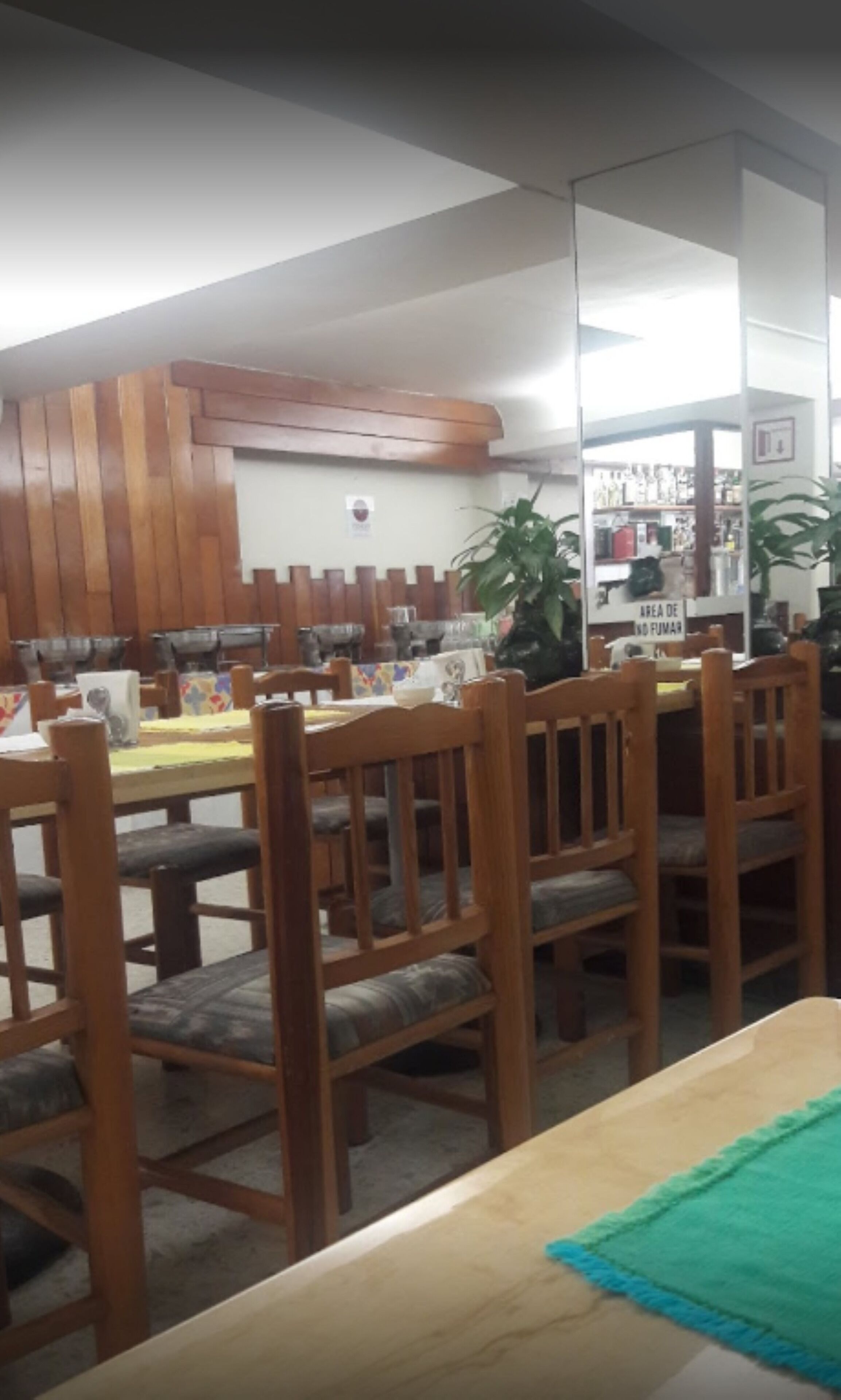 Un comedor con mesas y sillas de madera, una alfombra verde y un estante en la pared con varios objetos.