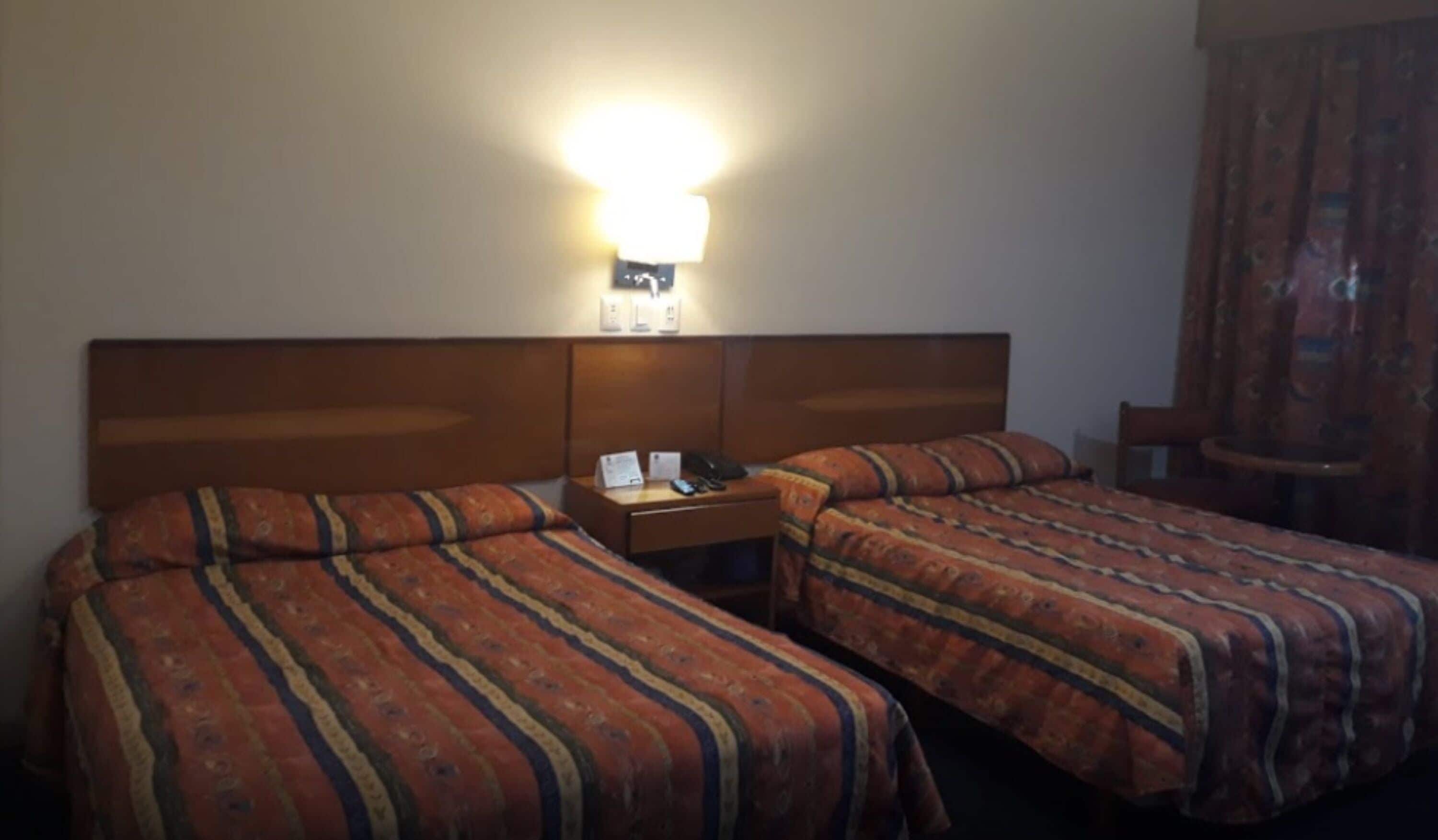 A hotel room with two beds, a nightstand with a lamp, and a chair.