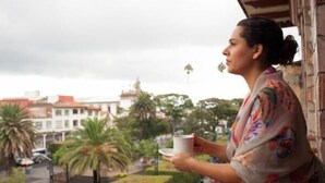 Una persona sosteniendo una taza, con vistas a un paisaje urbano con edificios y palmeras.
