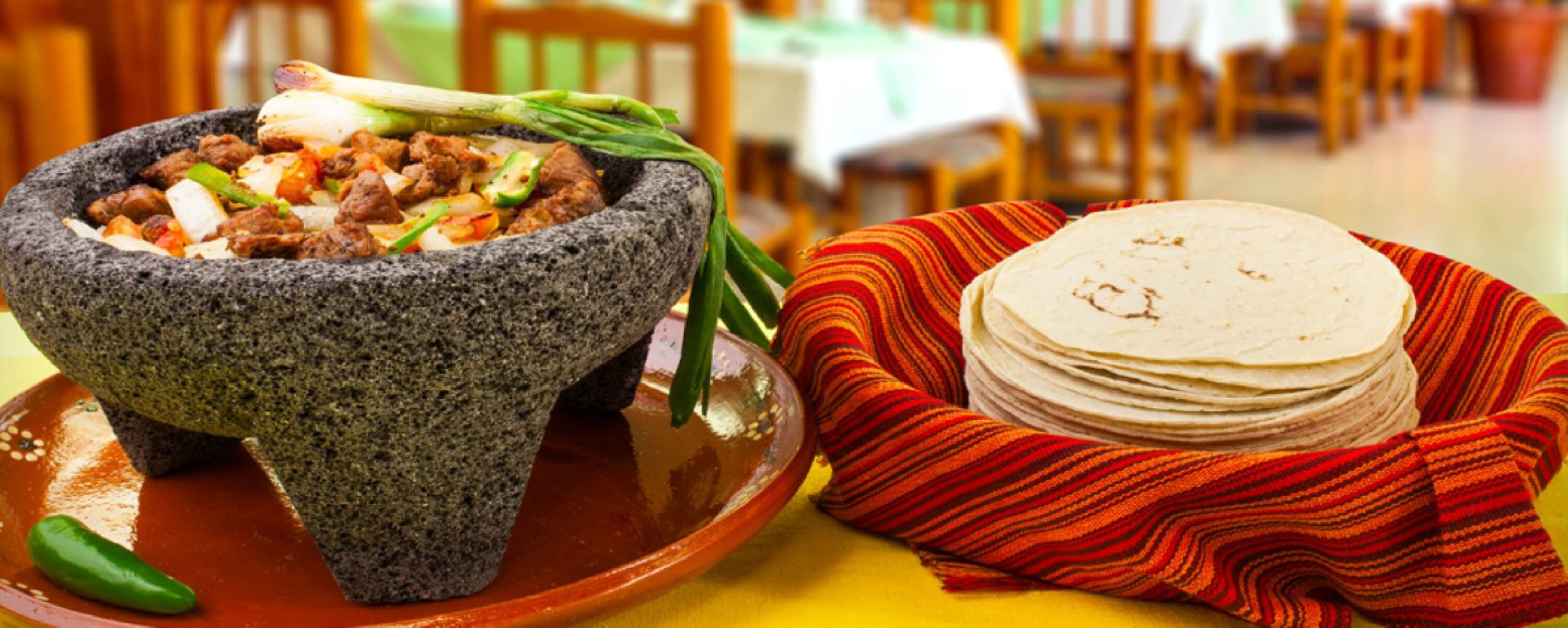 A stone mortar with food, a stack of tortillas, and a woven cloth on a table.