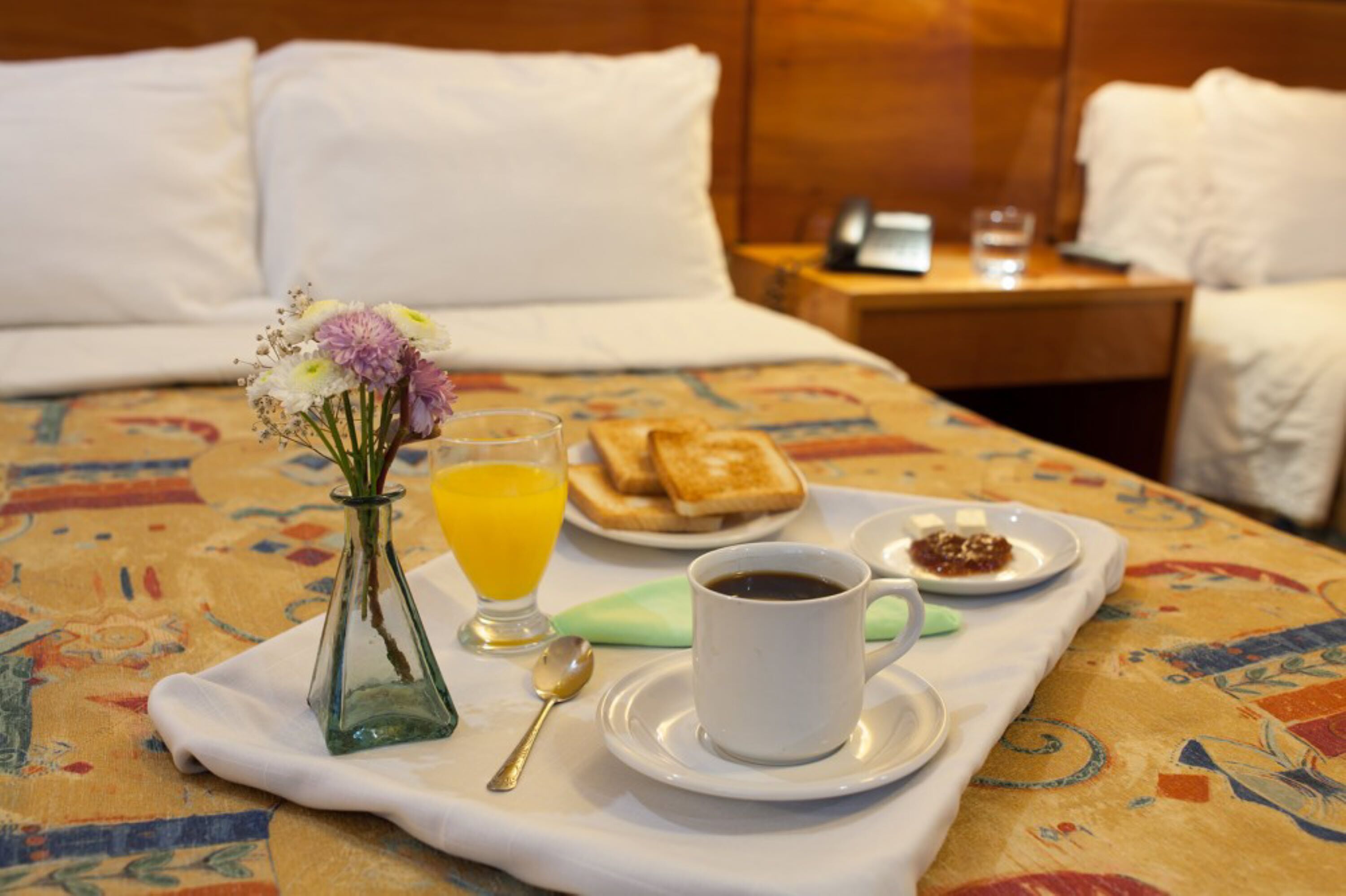 A neatly made breakfast tray on a bed with toast, a cup of coffee, a glass of orange juice, and a small vase with flowers.