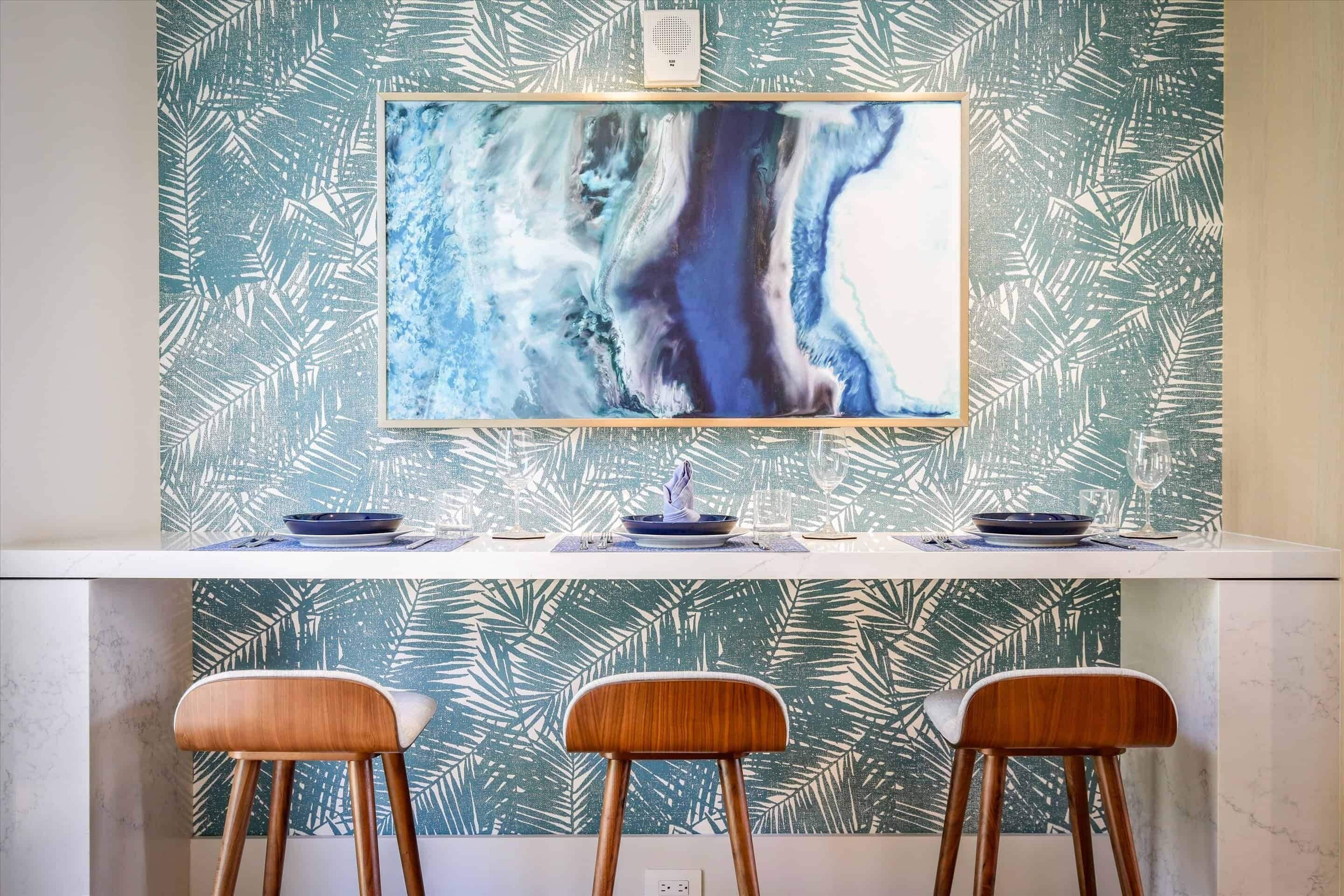 A kitchen with a large abstract painting, a white countertop, and three wooden bar stools.