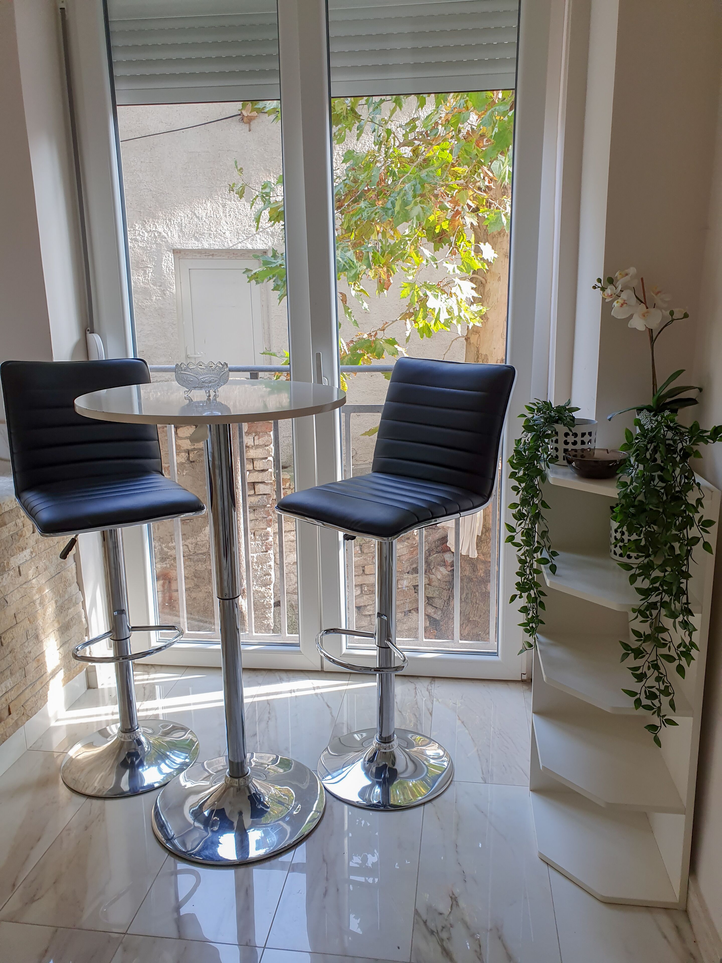 Ein modernes Zimmer mit zwei schwarzen Barhocker, einem Glastisch und Blick auf einen Garten durch große Fenster.