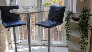 A modern room with two black bar stools, a glass table, and a view of a garden through large windows. - Srecko Centar Apartment (Belgrade)
