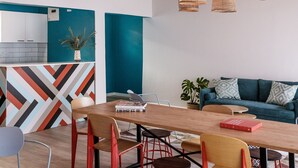 A modern living room with a wooden dining table, red and yellow chairs, a teal sofa, and wicker pendant lights.