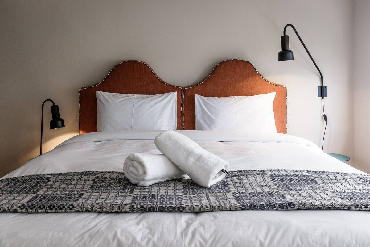 A neatly made bed with white linens, a rolled white towel, and a patterned throw blanket.