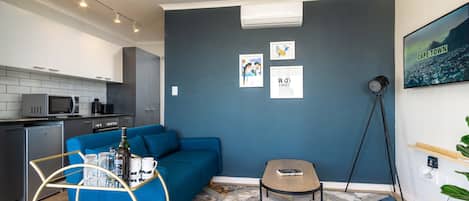 Una sala moderna con un sofá azul, una mesa de centro y un tapete estampado. Hay una zona de cocina con microondas y refrigerador.