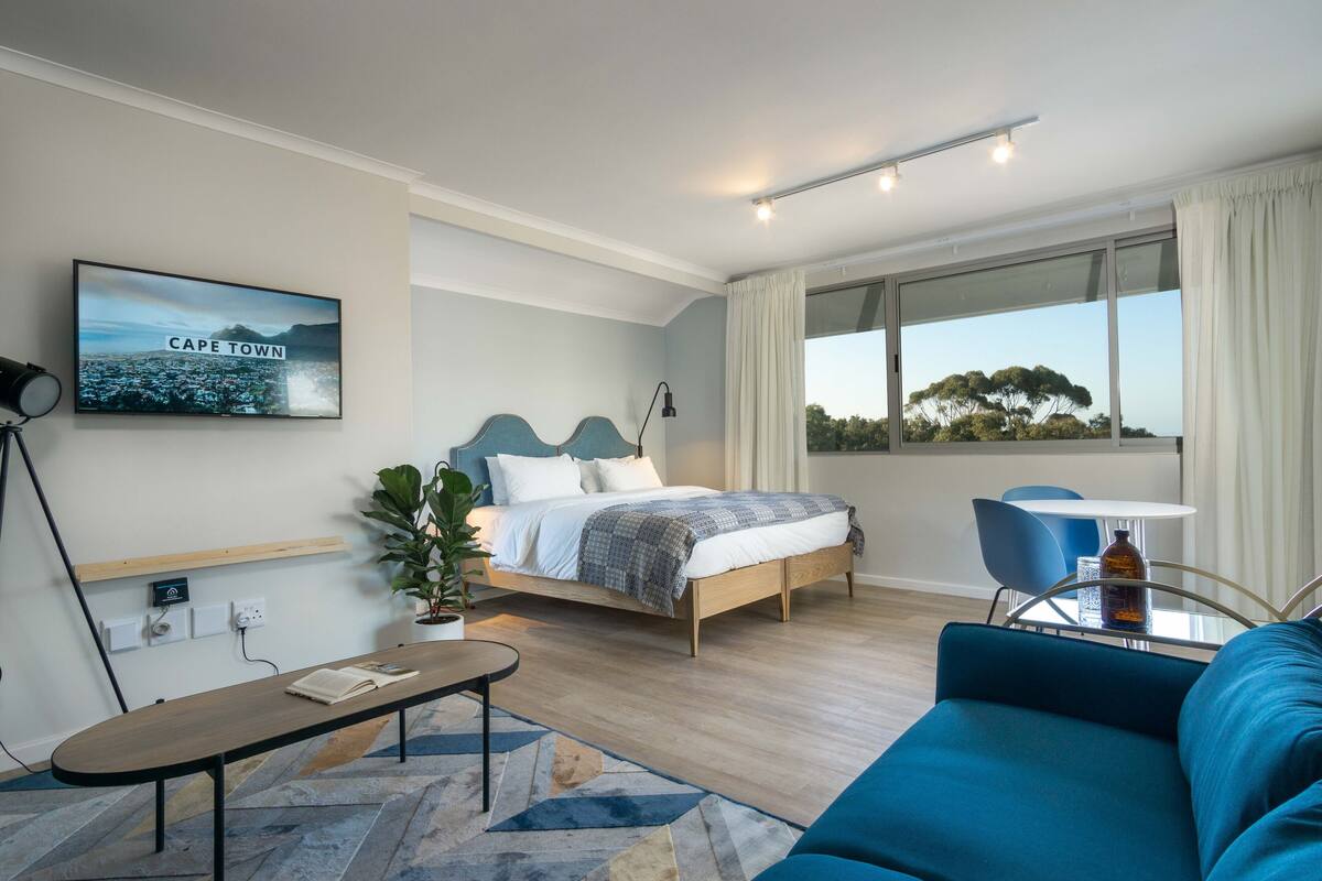 A modern hotel room with a bed, a desk, a sofa, a large window with a view, and a painting of Cape Town.