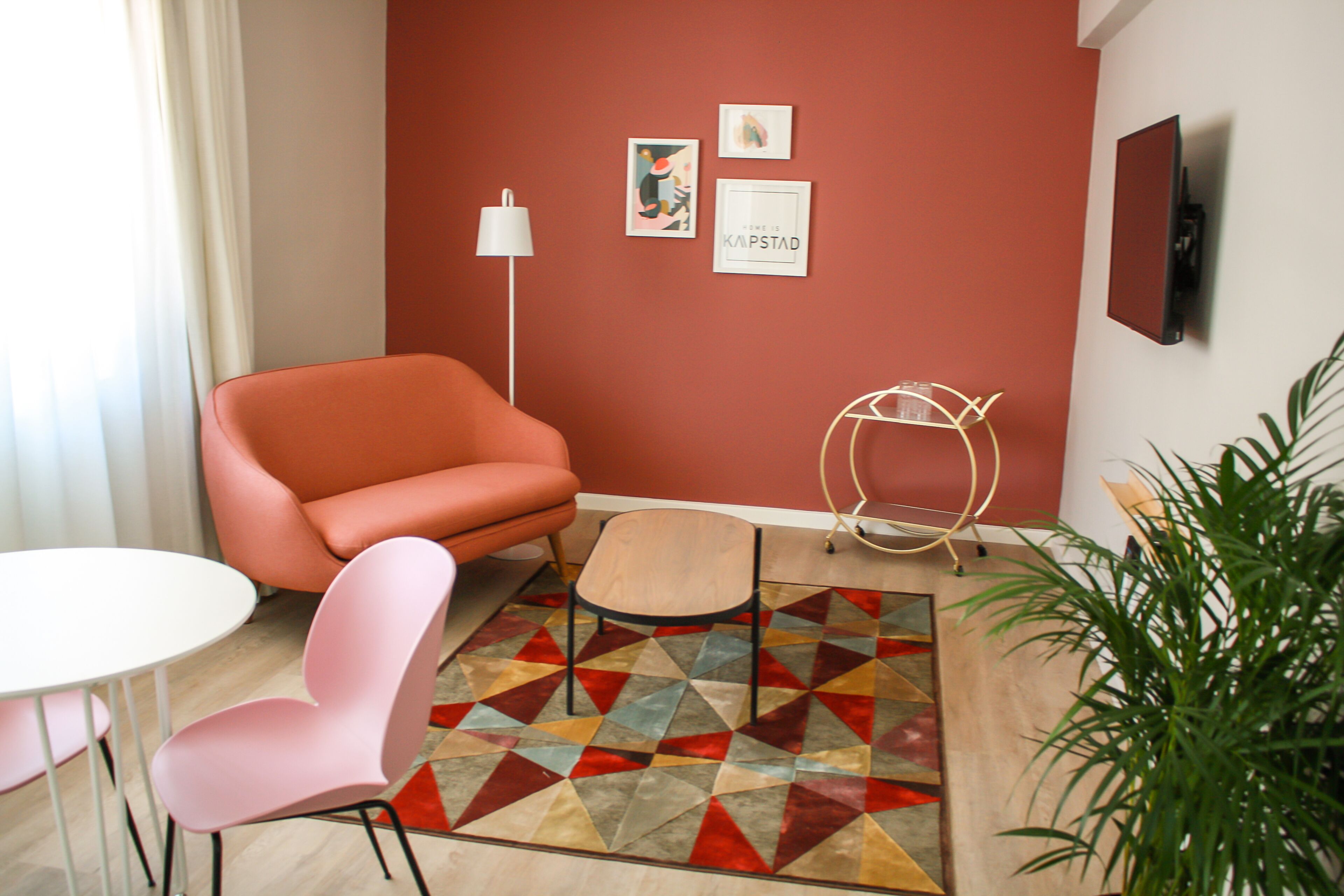 A modern room with a red accent wall, a white round table, a pink chair, a small wooden stool, and a geometric patterned rug.