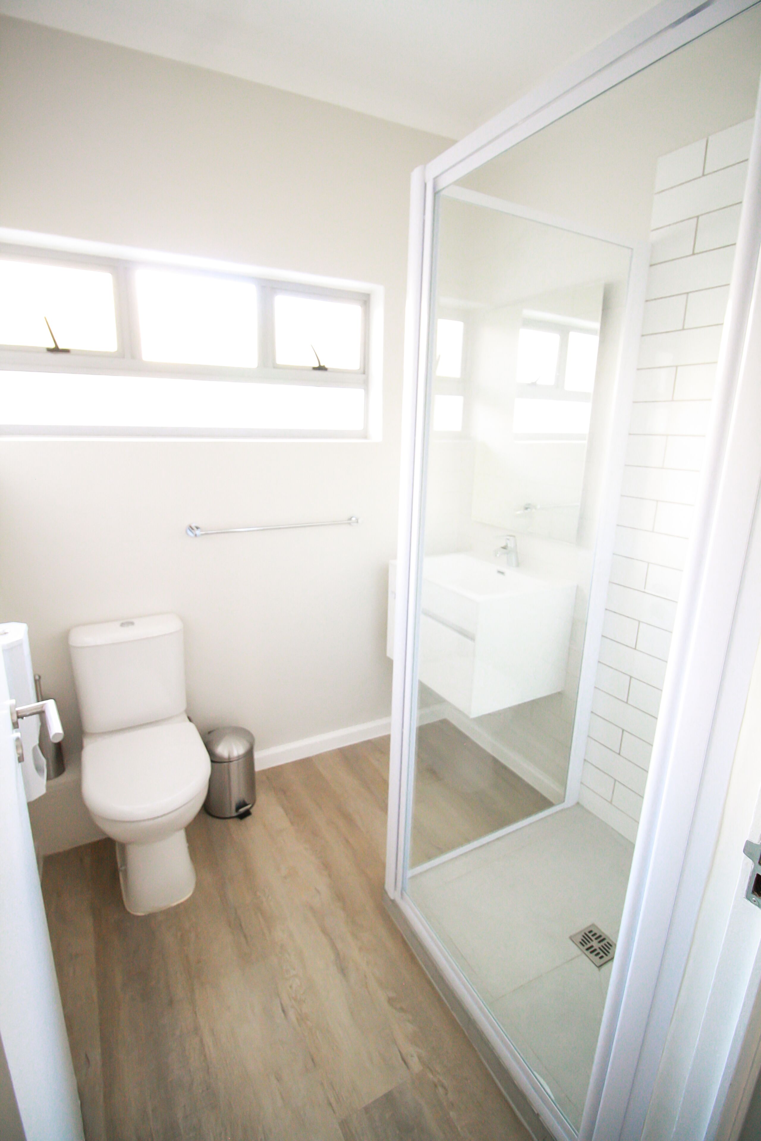 Une salle de bain moderne équipée de toilettes, d’une poubelle, d’une douche à parois de verre et d’une fenêtre située au-dessus des toilettes.